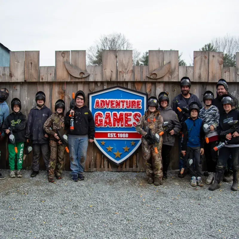 Group of people, some with paintball guns, pose in front of an Adventure Games sign.