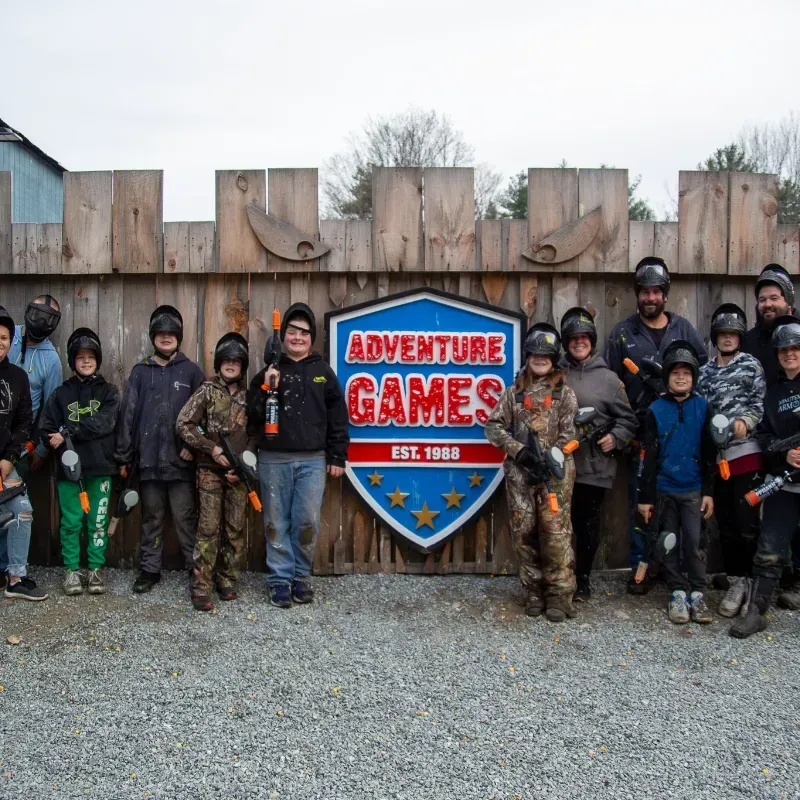 Group of people in paintball gear pose in front of Adventure Games sign.