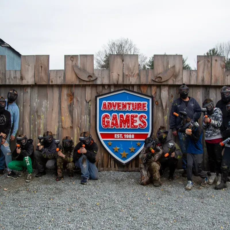 People in paintball gear posing in front of Adventure Games sign and wooden fence.