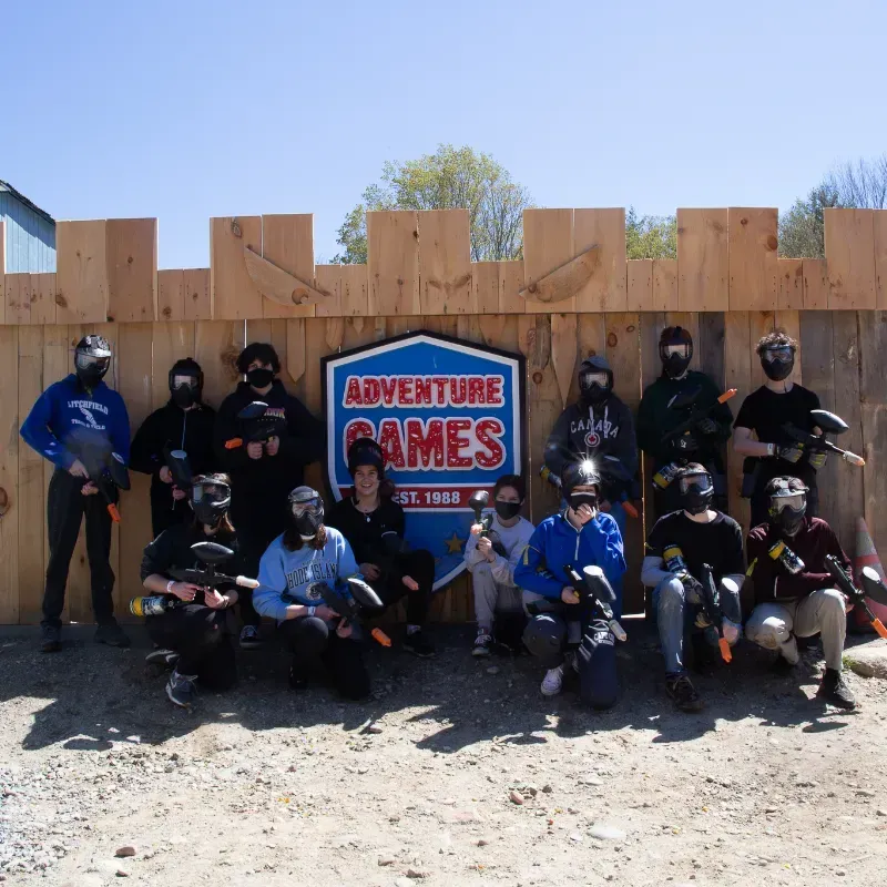 Group poses in front of Adventure Games sign, holding paintball guns, wearing masks, outside on a sunny day.