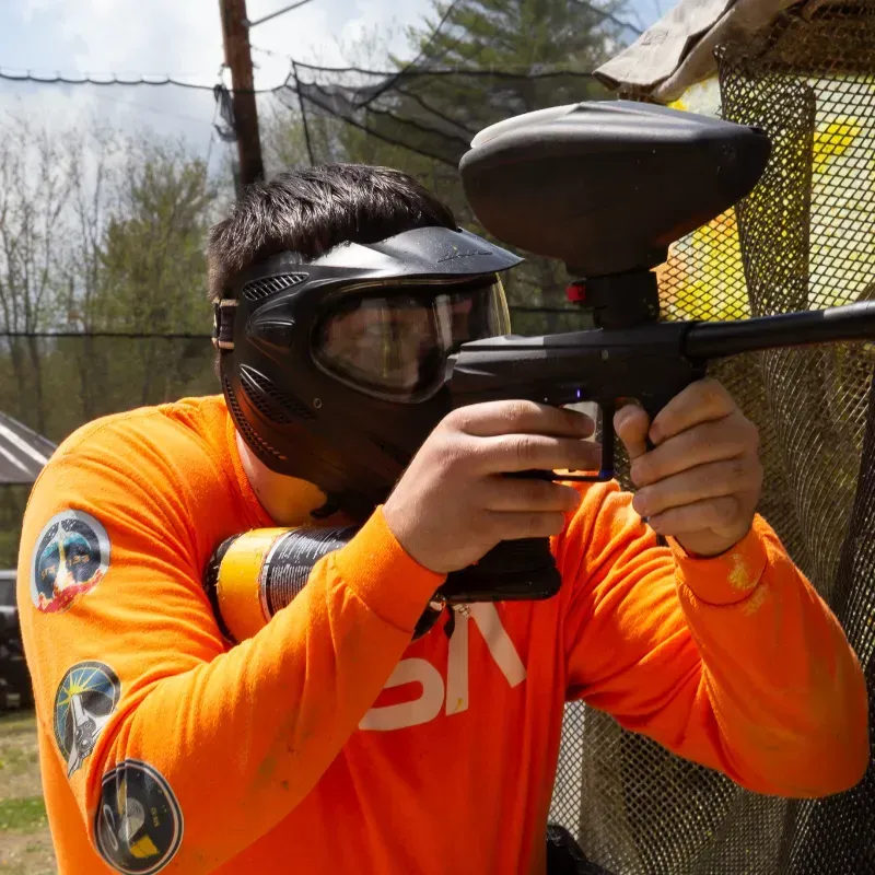 A person in orange shirt and paintball mask aims a paintball gun outdoors.