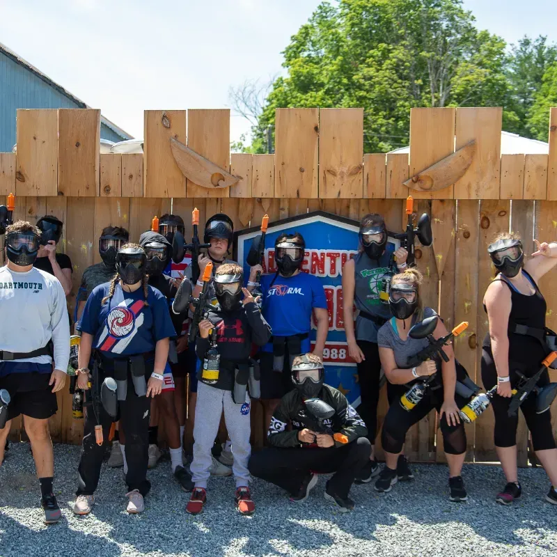 Group of people with paintball gear in front of a wooden fort. Some are holding guns.