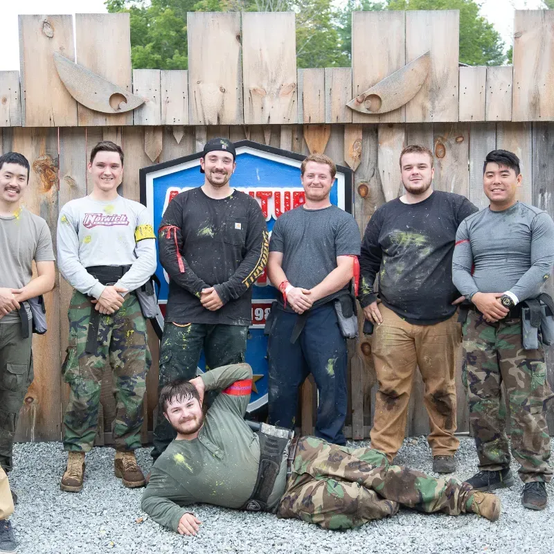 Group of men in paintball gear, posing outside. One man lies on the ground, smiling.