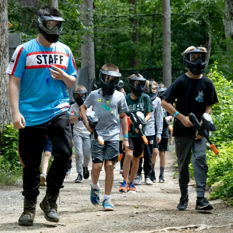 A group of children and a staff member walk along a path, geared up for paintball.
