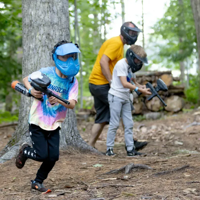 Children and an adult playing paintball in a forest, wearing masks and holding paintball guns.