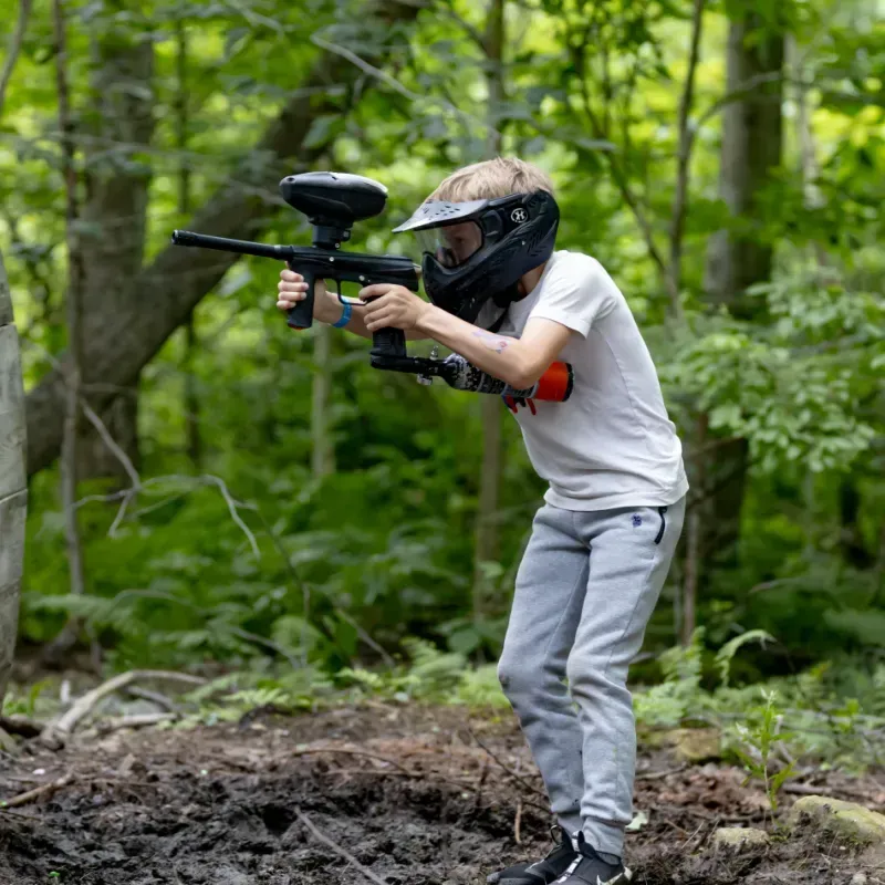 A person in a paintball helmet aims a paintball gun in a wooded area.