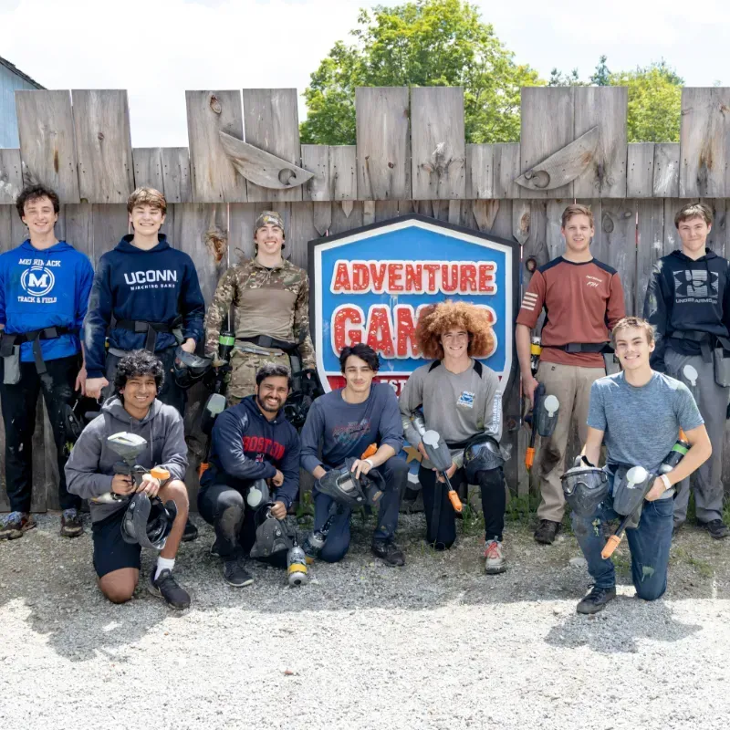 A group of young men pose at an adventure games location, holding paintball guns and smiling.