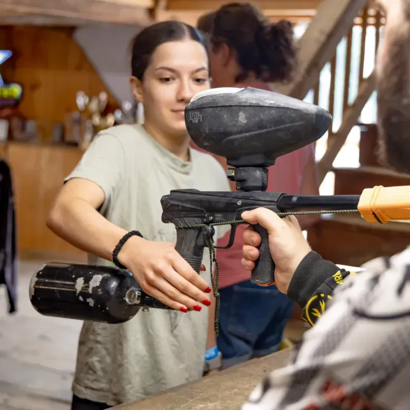 Woman with ponytail adjusts paintball gun for a person wearing checkered shirt, inside a building.