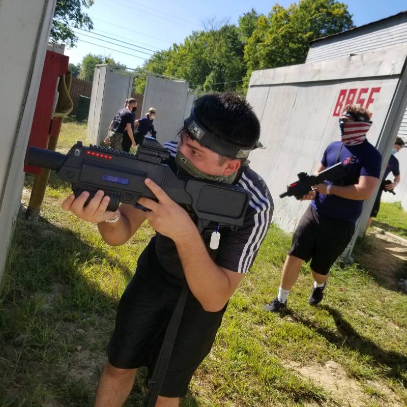 Teenagers playing laser tag outdoors, aiming laser guns. One wears a black Adidas shirt and mask.