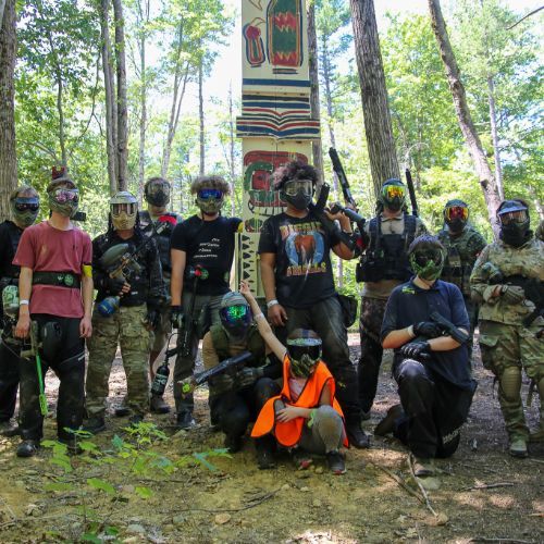 Group of people in paintball gear posing in a wooded area, some smiling, near a wooden structure.