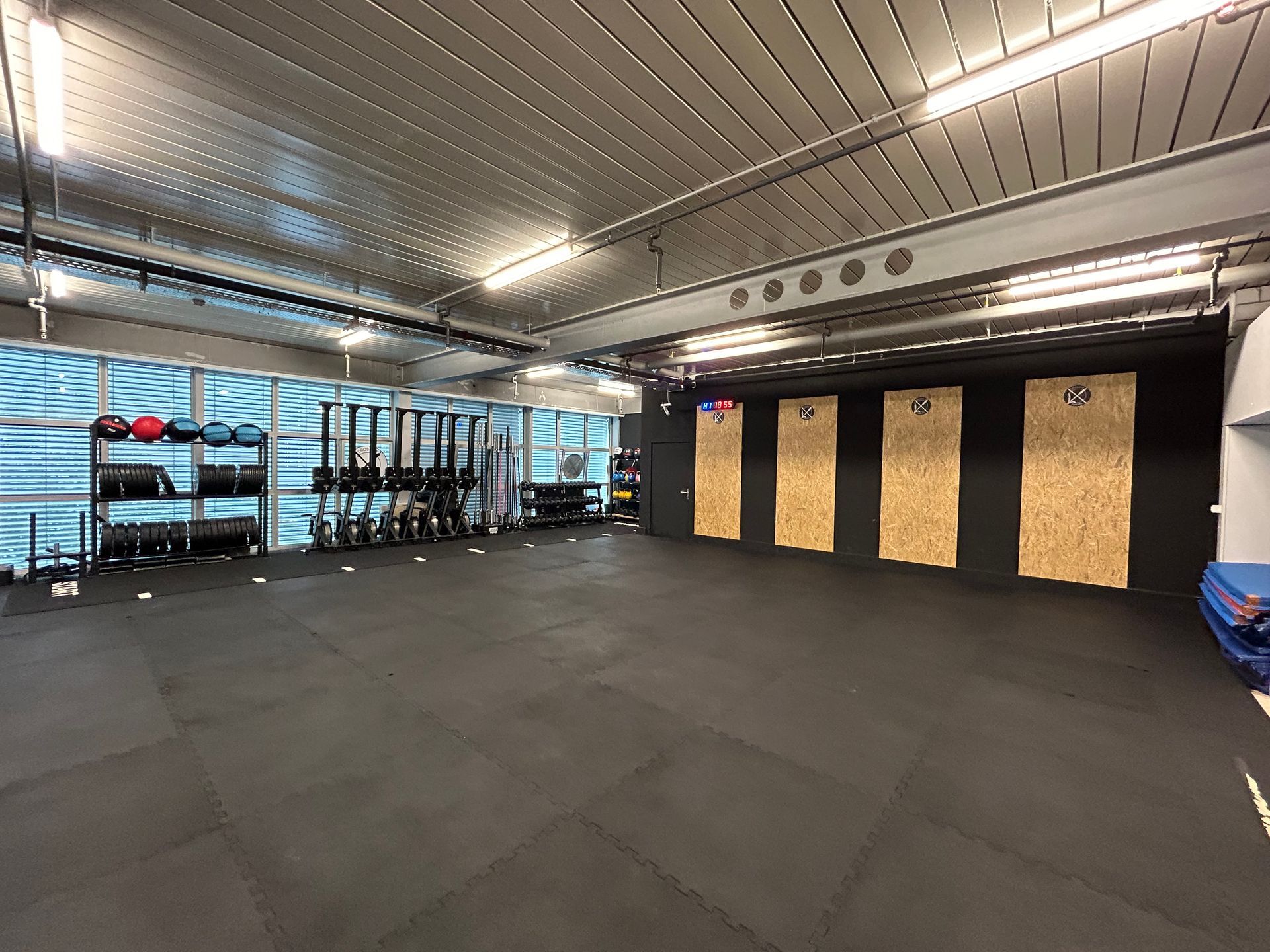 Empty gym studio in Thalwil with black rubber floor, wall-mounted wooden panels, rowing machines, and fitness equipment