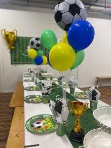 A table decorated for a soccer-themed party with balloon centerpieces, trophy accents, and themed plates and cups.