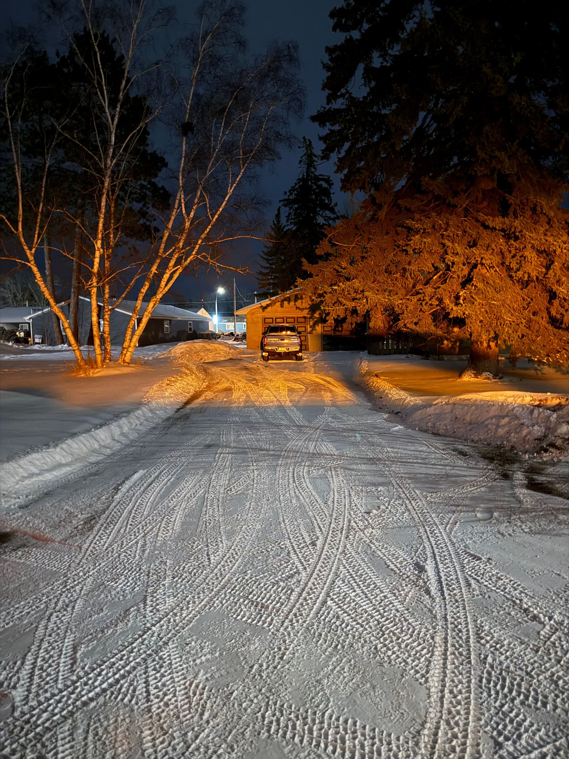 A snowy driveway at night, leading to a garage with a car parked inside, flanked by trees lit by warm outdoor lights.