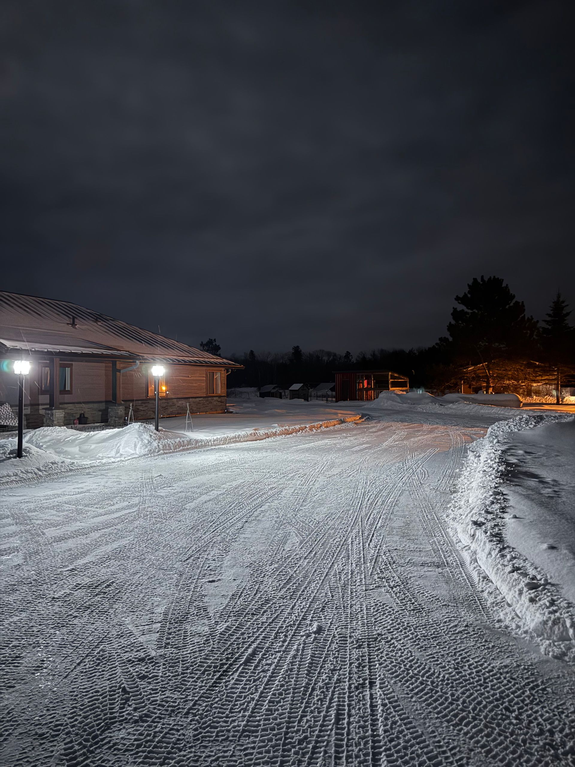 A snow-covered driveway at night, lit by two outdoor lamps in front of a house, under a dark, cloudy sky.