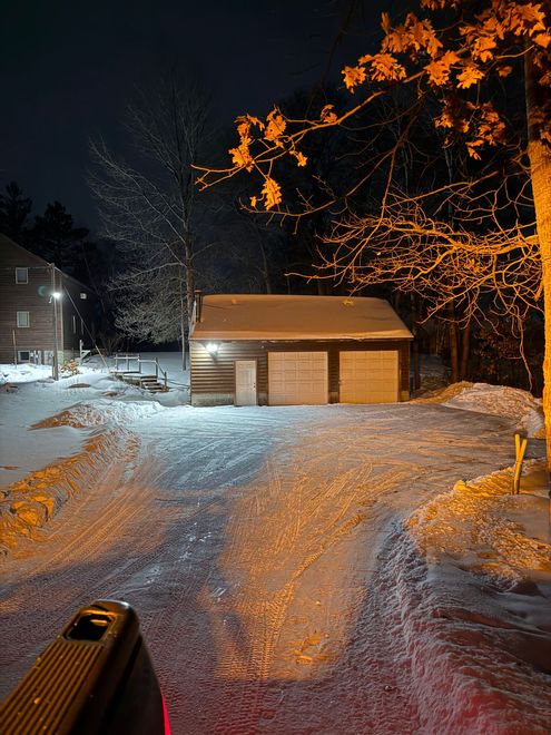 A nighttime view from a vehicle looking at a detached two-car garage covered in snow, illuminated by bright floodlights.