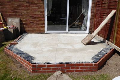 A new concrete patio slab enclosed by red brick retaining walls sits outside a glass sliding door.