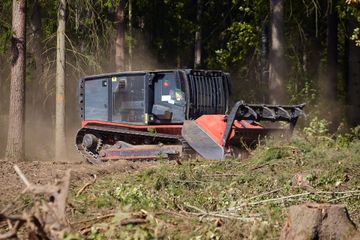 A red track-mounted forestry mulcher clearing brush in a forest, kicking up dust while operating among trees.