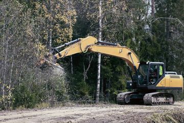 A yellow excavator with a mulcher attachment clearing brush and small trees along the edge of a forest road.