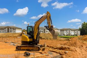 A yellow excavator sits in a construction site with dirt mounds and apartment buildings in the background under a blue sky.