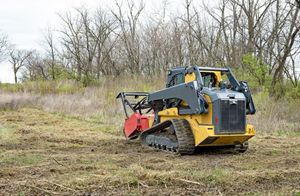 A yellow compact track loader with a mulching attachment clears brush in a field under a cloudy sky.