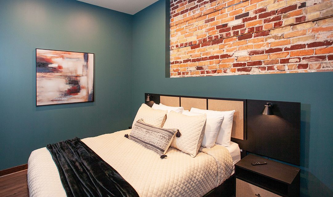Bedroom with teal walls, brick accent, and a neatly made bed with white bedding and dark throw blanket
