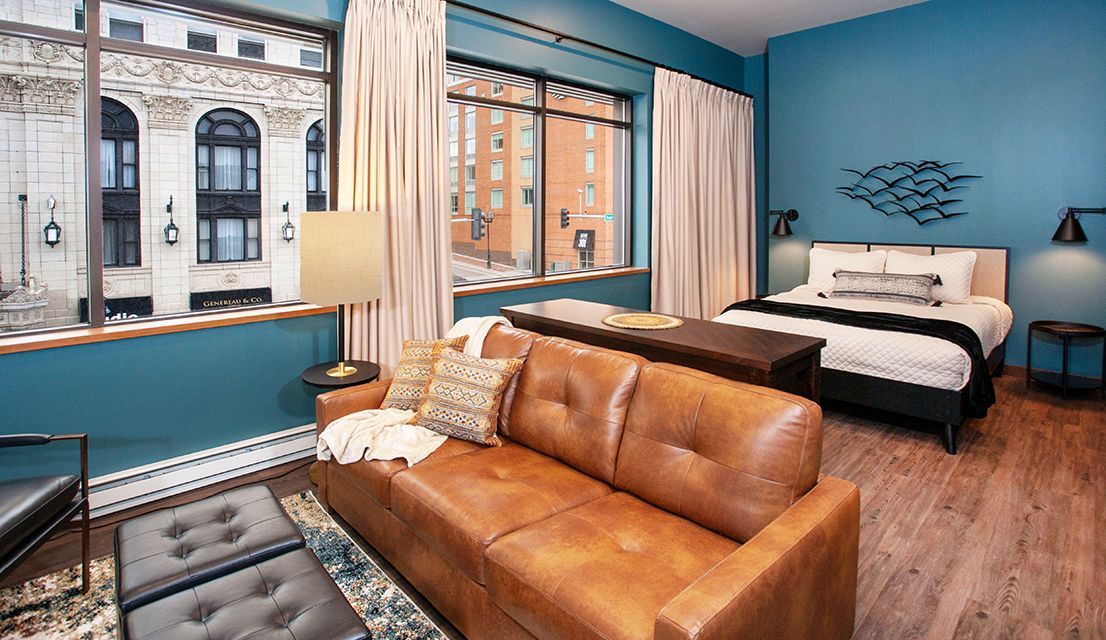 Cozy teal bedroom with a brown leather sofa, bed, and large windows overlooking brick buildings