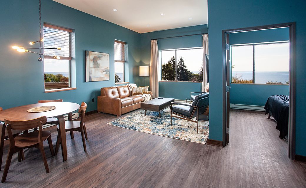 Bright blue living room with sofa, chairs, rugs, and open doorway to a bedroom.