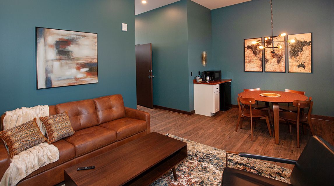 Modern living room with teal walls, brown leather sofa, small dining table, and abstract wall art.