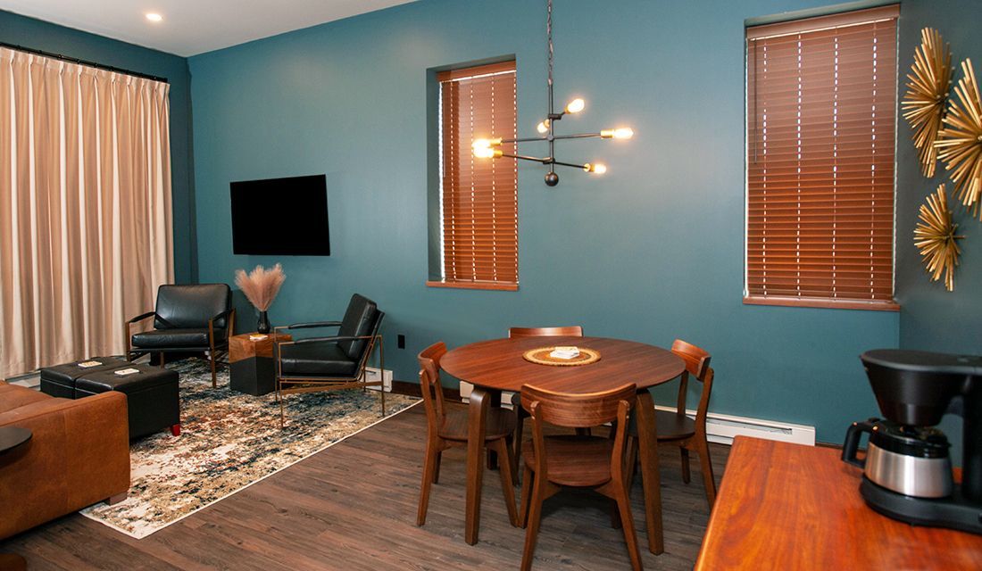 Modern living room with teal walls, round wooden table, chairs, rug, windows, and wall-mounted TV