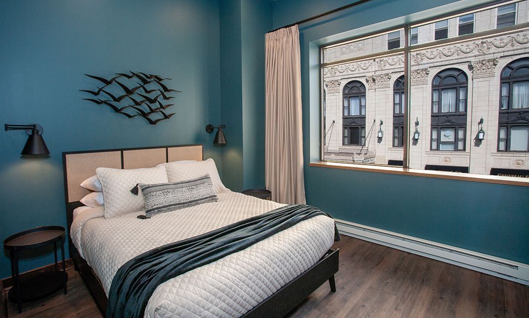 Bright blue bedroom with a bed, wall art, and a large window overlooking buildings
