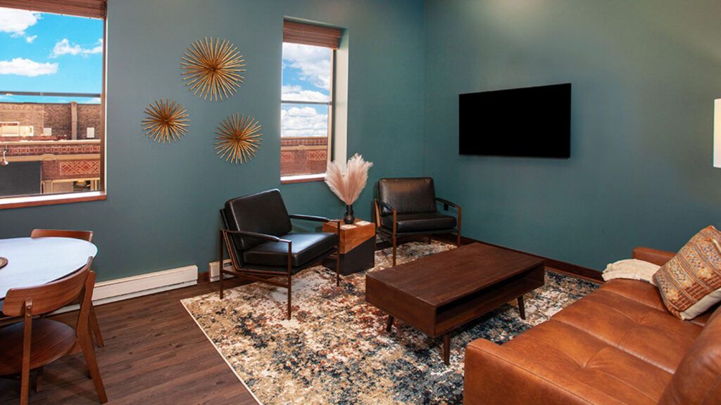 Modern living room with teal walls, leather chairs, rug, coffee table, wall art, and mounted TV.