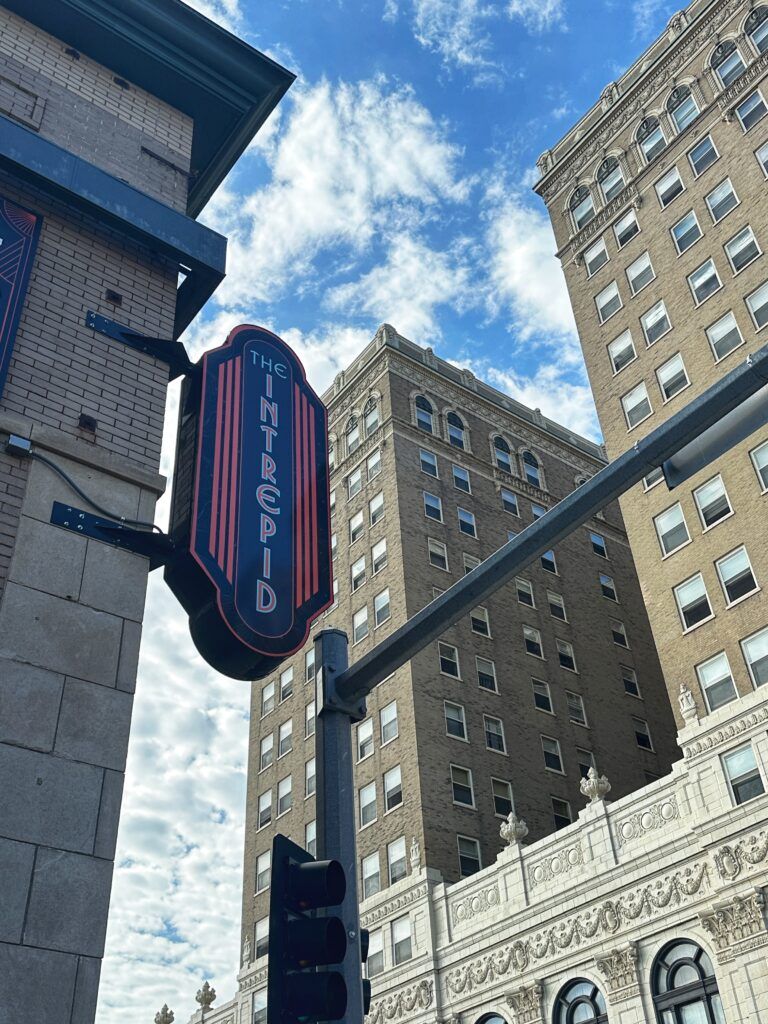 Downtown city street with tall brick buildings and a vertical theater sign against a cloudy blue sky