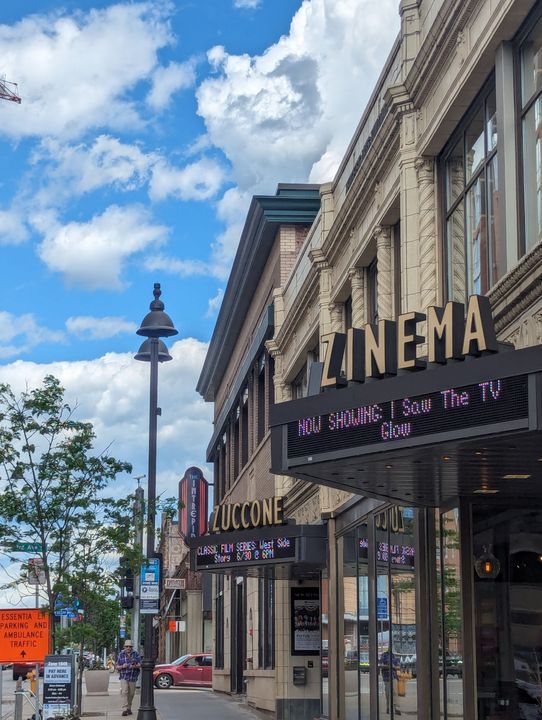 Downtown street with storefronts, including a cinema marquee, under a bright cloudy sky