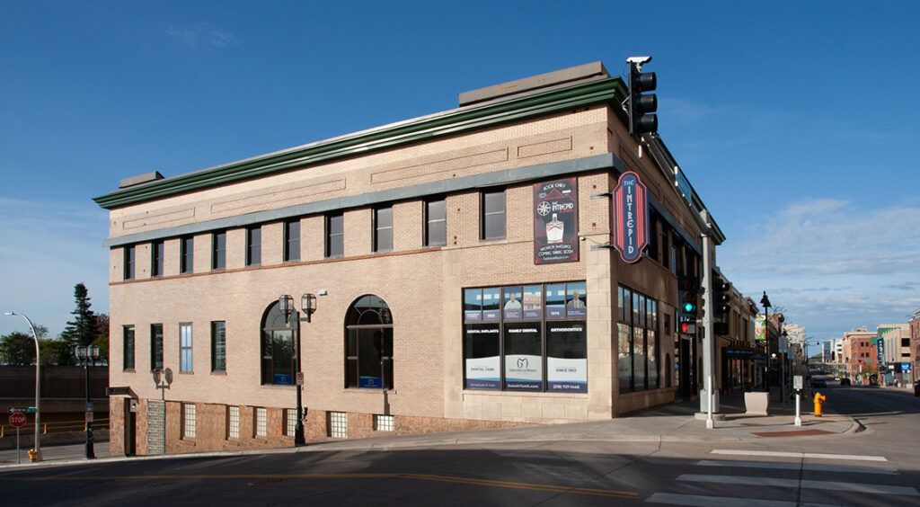 Corner view of a tan brick building with arched windows and a green roof on a city street.