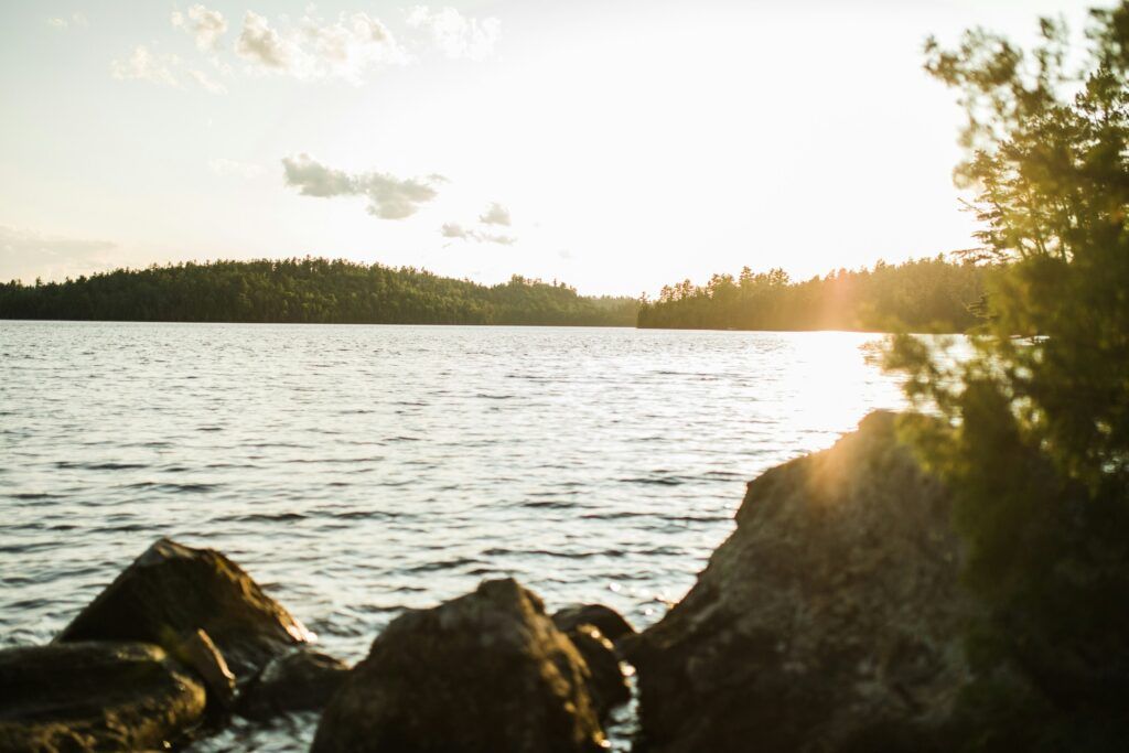 Sunlit lake with rocky shoreline and distant tree-lined shore at sunset
