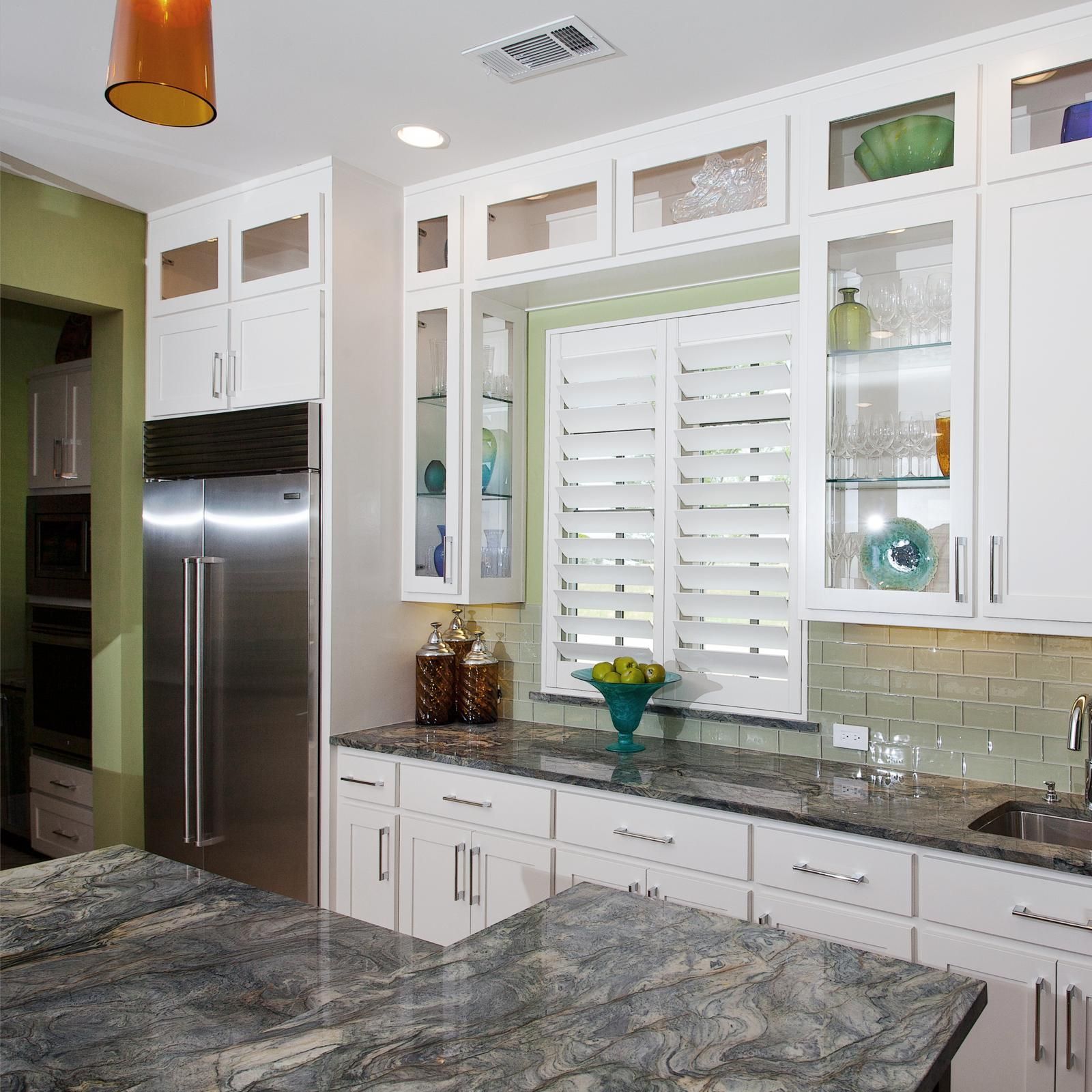 A modern kitchen featuring white cabinets, stainless steel refrigerator, granite countertops, and a window with shutters.