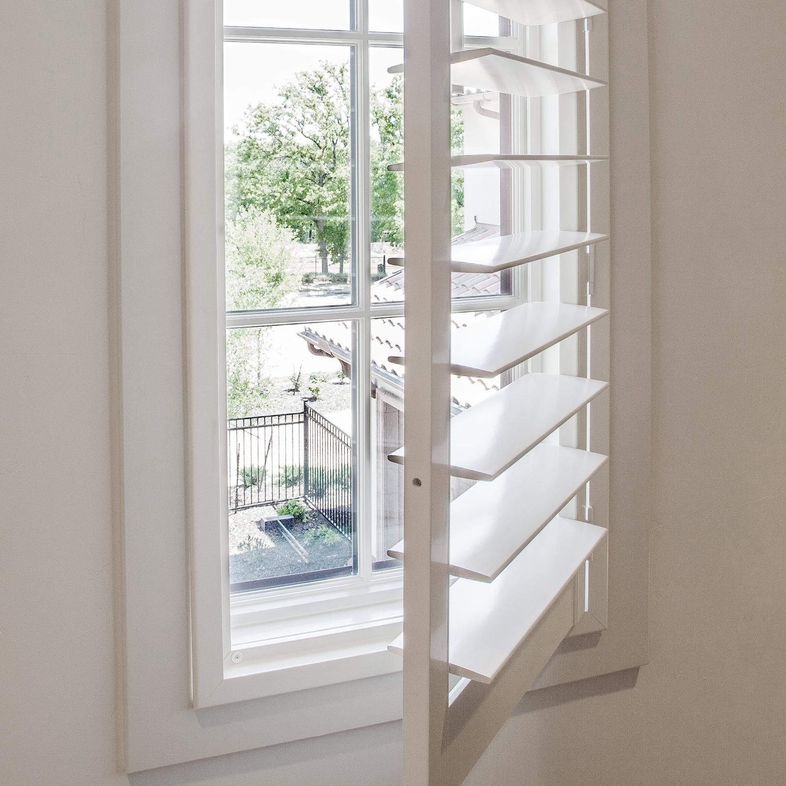 White wooden plantation shutters partially open in front of a window, looking out onto a bright green outdoor scene.