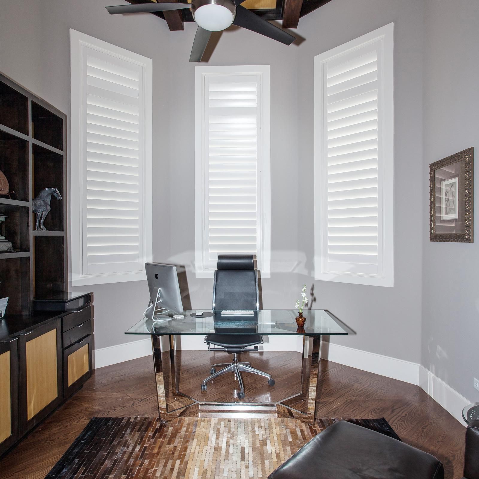A home office featuring a glass desk, black chair, and large windows with white shutters in a neutral-toned room.