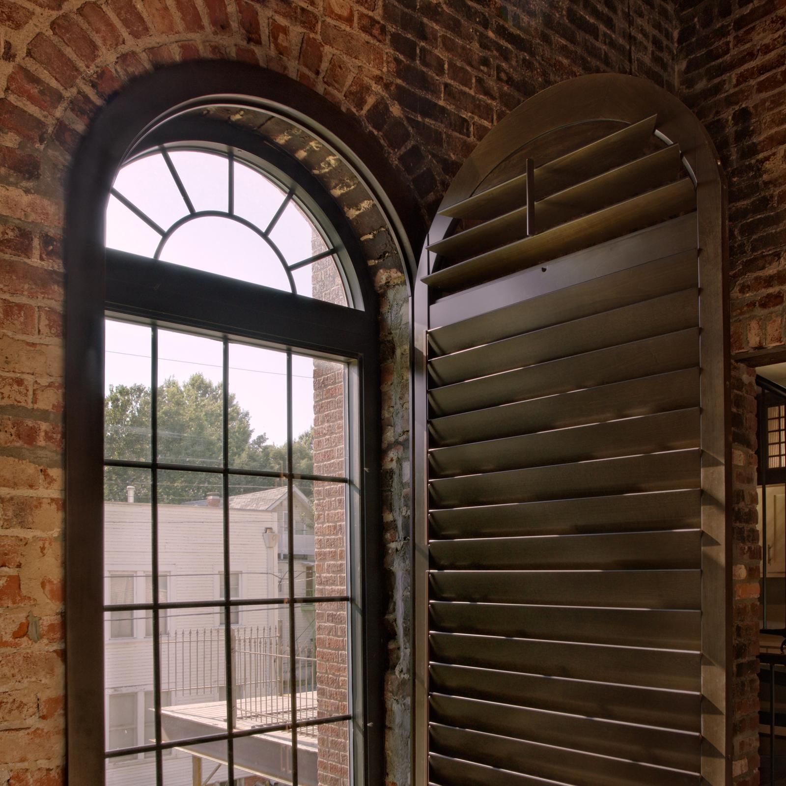 An arched window with a grid pane is set in an exposed brick wall, partially covered by a tall, closed wooden shutter.