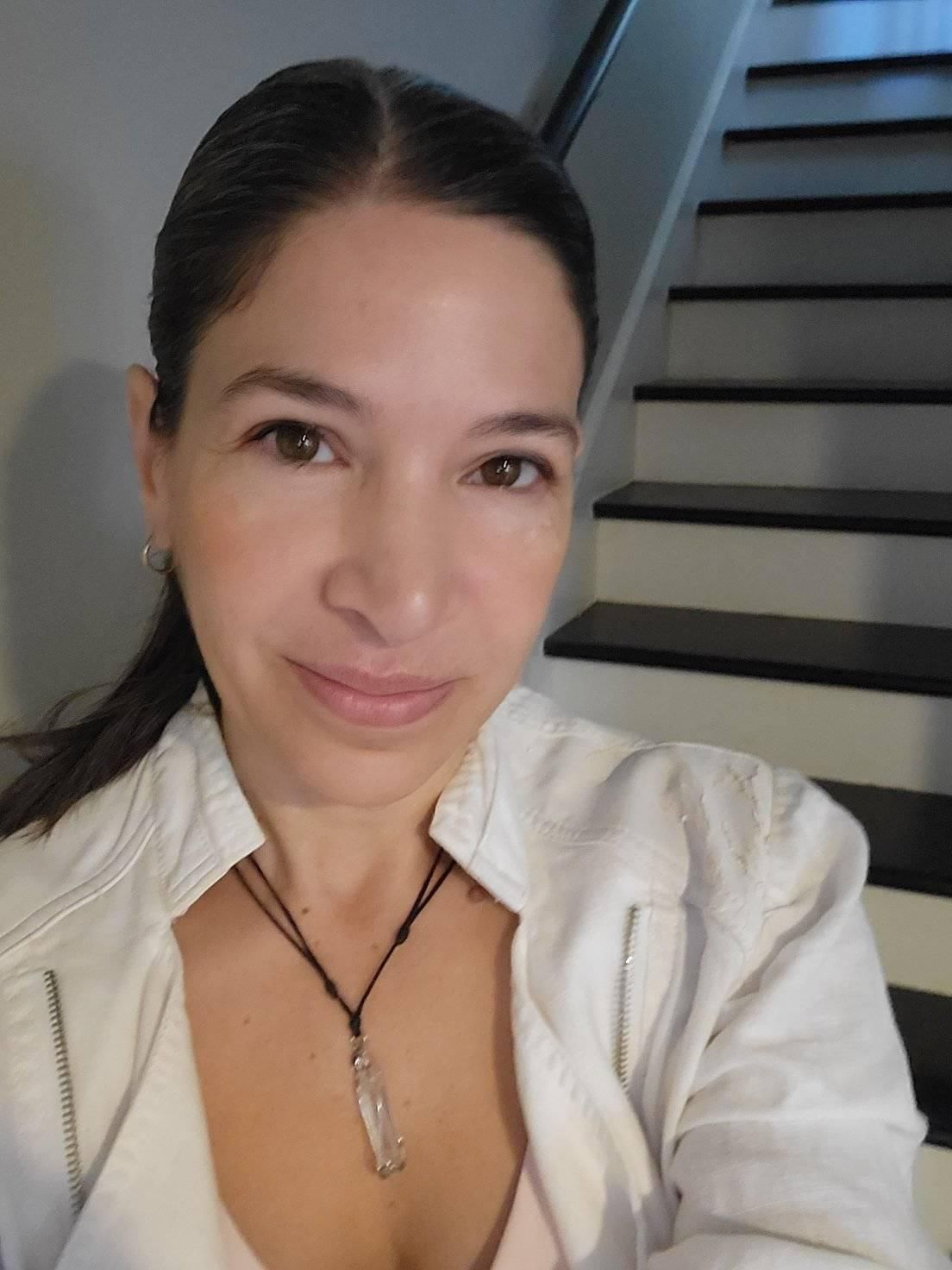 Woman in white jacket smiles, standing near stairs. She wears a necklace.