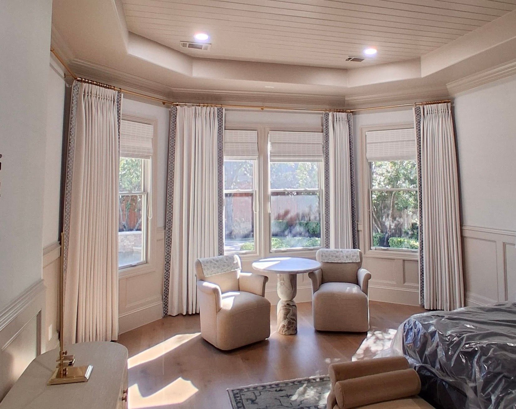 A light-filled seating area with two upholstered chairs, a small table, floor-to-ceiling cream curtains, and tray ceiling.