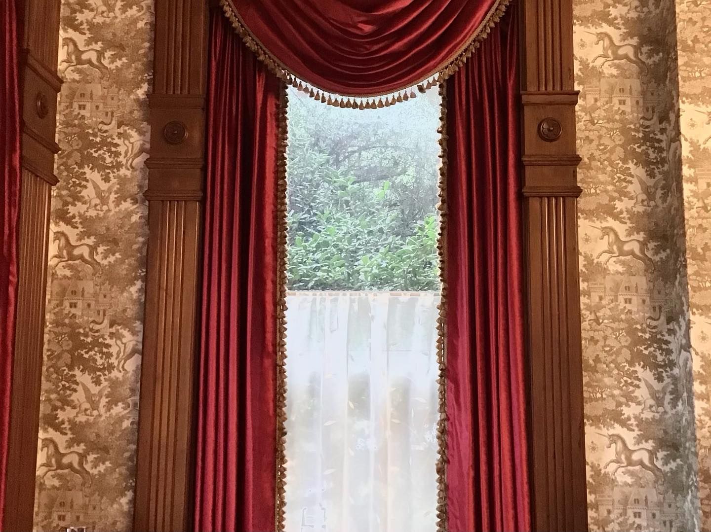 Red velvet curtains frame a window, with ornate wooden trim and patterned wallpaper.