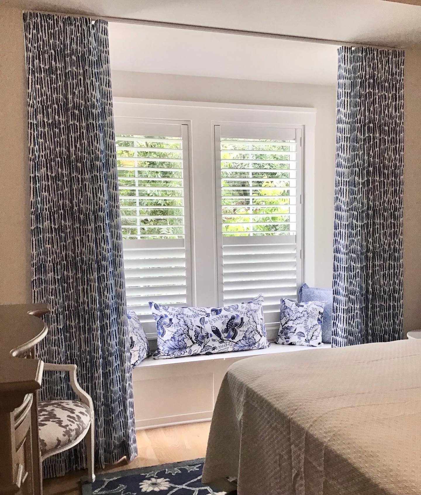 Bedroom window with blue patterned curtains, shutters, and decorative pillows.