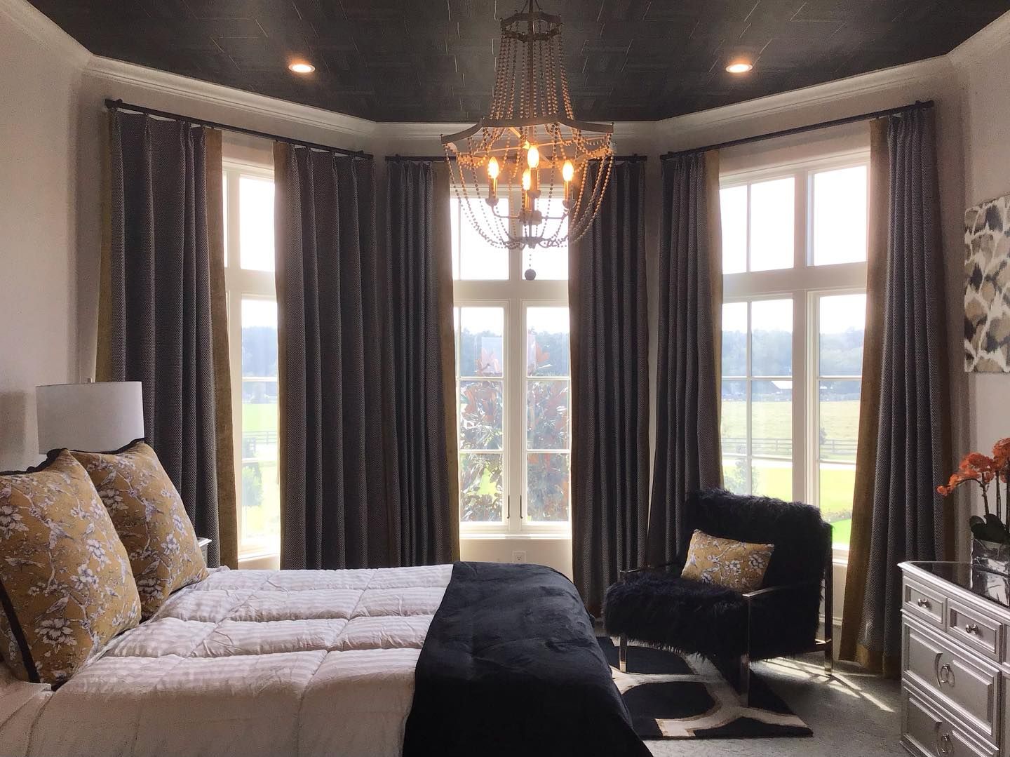 Bedroom with large windows, dark gray curtains, chandelier, bed with gold pillows, and a black armchair.
