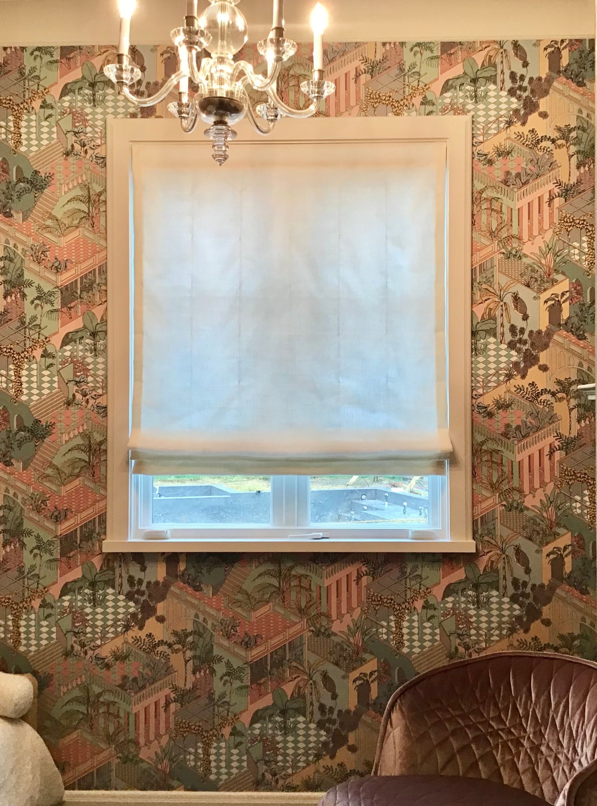 A window with a translucent white roller shade, framed by ornate, multi-colored patterned wallpaper and a chandelier.
