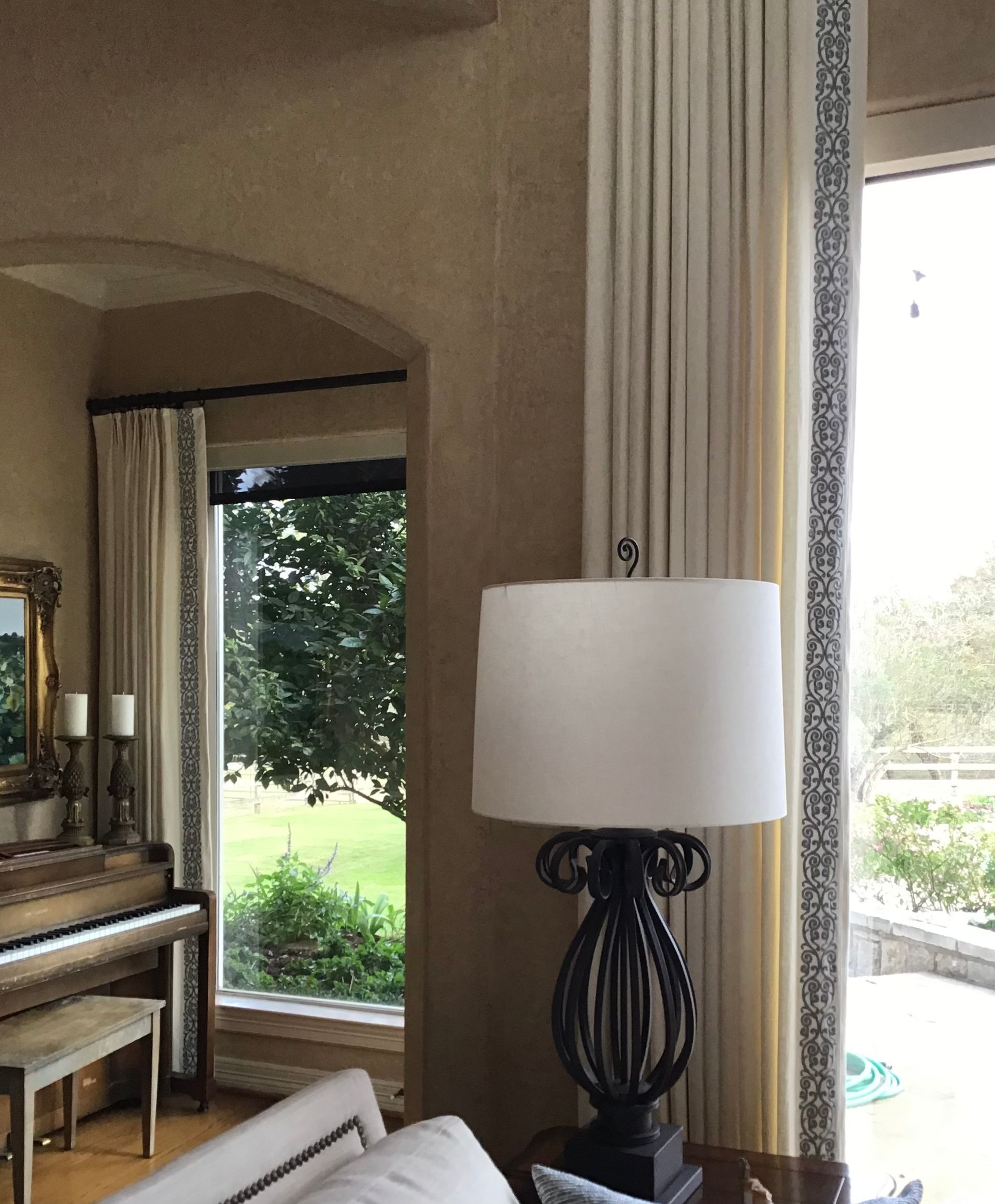A room with beige textured walls, a lamp, a piano, and cream-colored curtains with patterned blue borders by a window.