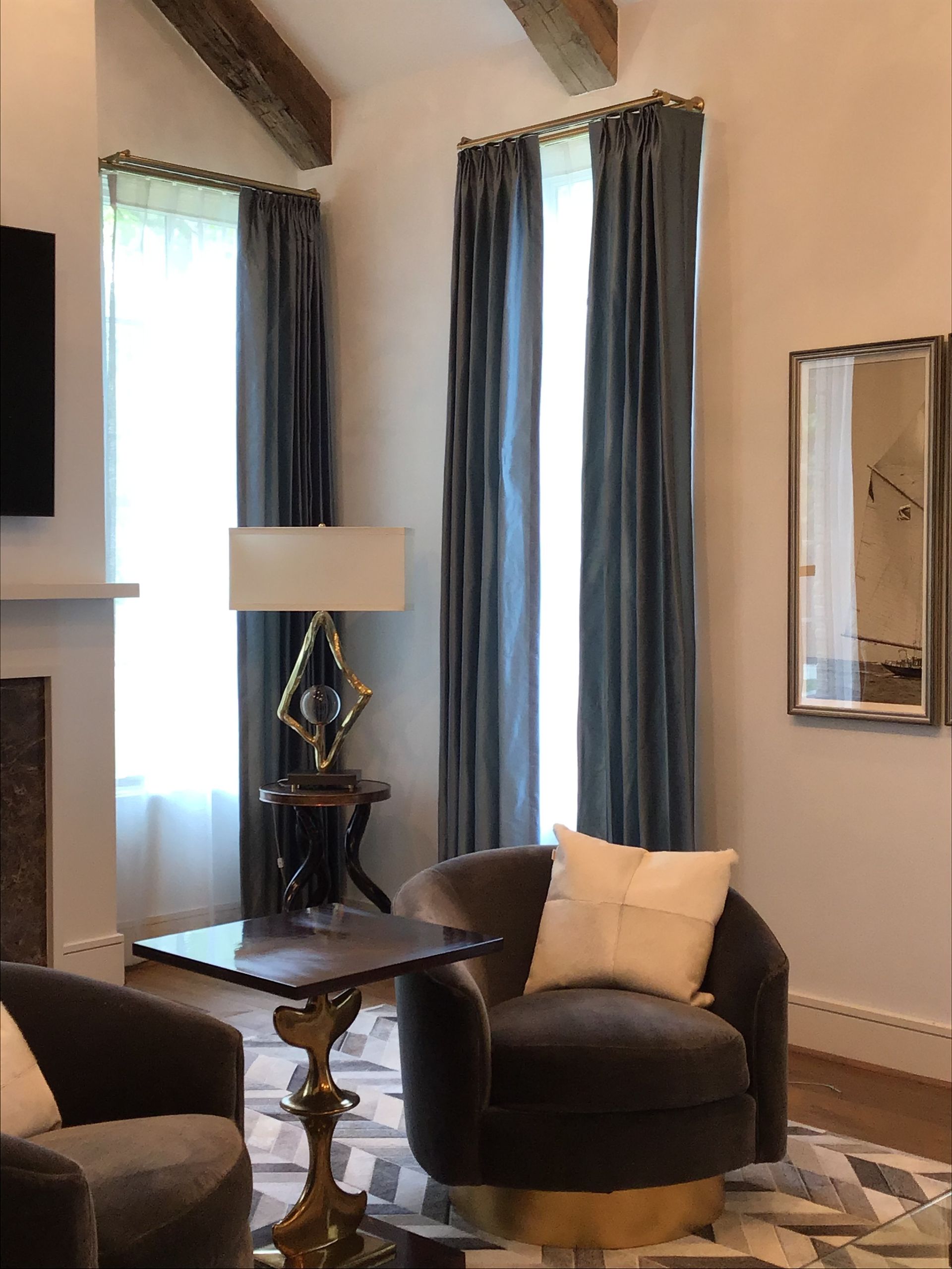 Two plush dark armchairs face a small table and a lamp in a living room with tall windows and blue velvet drapes.