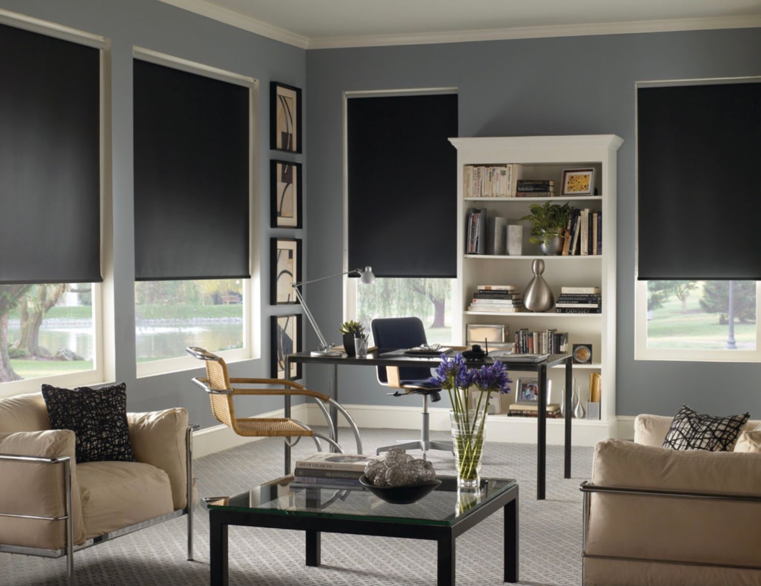 A home office with gray walls, black roller shades, two beige armchairs, a glass coffee table, and a white bookcase.