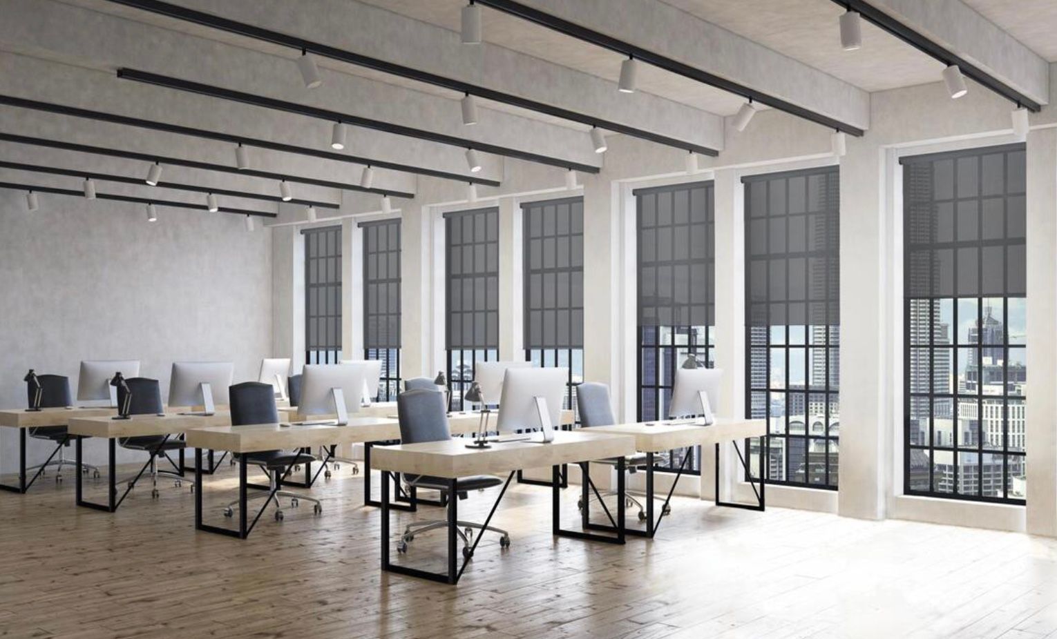 A modern, open-plan office with desks, computers, and large windows with dark roller shades, viewed from an angle.