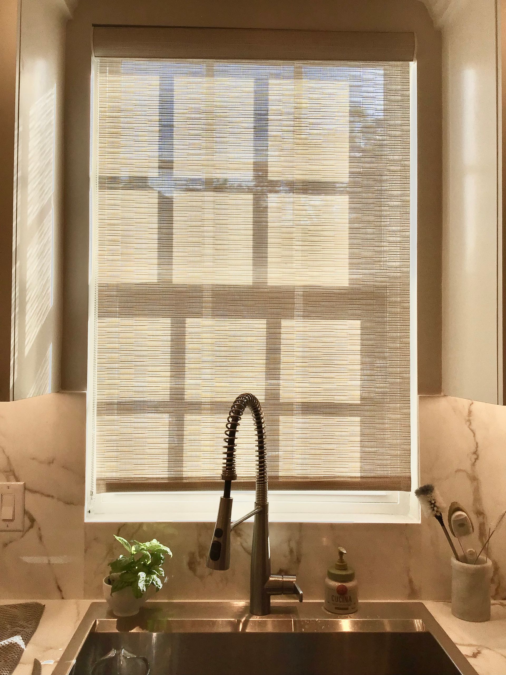 A kitchen sink with a faucet and a small potted plant in front of a window covered by a textured light brown bamboo shade.